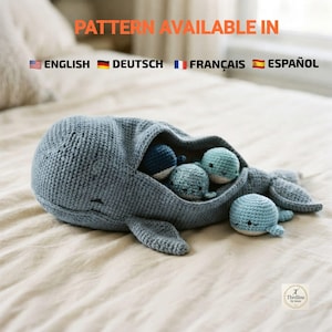 May include: A large, blue, crocheted whale toy with an open mouth containing three smaller, round, crocheted whale toys in shades of blue and teal. The large whale has a textured, yarn-based surface and a tail fin extending to the right side.