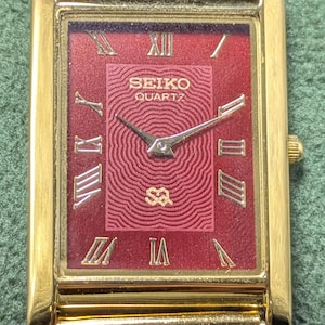 May include: A rectangular gold-tone Seiko Quartz watch with a deep red dial featuring a concentric wave pattern, gold Roman numeral hour markers, and a gold-tone mesh strap. The dial displays the text SEIKO QUARTZ and a small SQ logo.