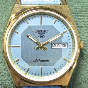 May include: A gold-tone Seiko 5 automatic watch with a light blue dial featuring a central grey octagon shape, gold-tone hands and hour markers, and a day-date window at the three o'clock position. The watch has a light blue textured leather strap.