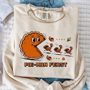 Könnte beinhalten: Ein cremefarbenes Sweatshirt mit einer Grafik einer orangefarbenen Tortenfigur, die drei Truthähne jagt. Das Design enthält Mais, Kürbisse und den Text PIE-MAN FEAST in Pixel-Schrift. Eine goldfarbene Kette liegt auf dem Kragen.