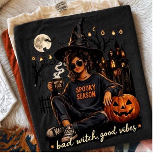 Könnte beinhalten: Ein schwarzes T-Shirt mit einer Grafik einer Person mit Hexenhut und Sonnenbrille, die eine Tasse mit der Aufschrift Witch Fuel hält. Das Shirt zeigt den Text Spooky Season und bad witch, good vibes, sowie einen Kürbis und ein Spukhaus.
