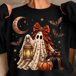 Könnte beinhalten: Ein schwarzes Sweatshirt mit einer Grafik eines Geistes, der in ein weißes Laken mit Kirsch- und Totenkopfmuster gehüllt ist und eine große orangefarbene Schleife trägt. Der Geist hält eine leuchtende Laterne neben orangefarbenen und schwarzen Kürbissen, umgeben von Fledermäusen, Sternen und einem Mond.
