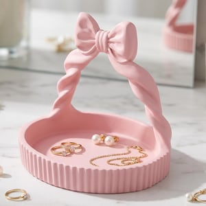 Può includere: Un vassoio portagioie circolare rosa chiaro con esterno a coste e un manico decorativo a forma di corda intrecciata sormontato da un fiocco. Il vassoio contiene due anelli dorati, un paio di orecchini di perle e una sottile catenina d'oro.