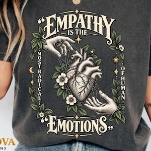 Può includere: Una t-shirt grigio antracite con una stampa grafica di un cuore anatomico tenuto da due mani, circondato da foglie e fiori. Il testo recita: "EMPATHY IS THE MOST RADICAL OF HUMAN EMOTIONS" in un carattere decorativo con accenti celestiali.