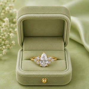 May include: A gold ring featuring a central pear-shaped clear gemstone flanked by three smaller round clear gemstones on each side. The ring is displayed inside an open, sage green velvet-lined jewelry box with a gold-toned metal clasp.