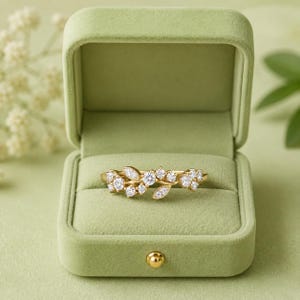 May include: A gold ring featuring a delicate branch-like design adorned with multiple small, round, clear, sparkling stones. The ring is displayed inside an open, light green, velvet-textured jewelry box with a single gold-toned metal button closure.