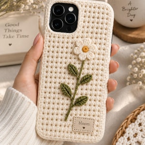 Può includere: Una custodia per telefono all'uncinetto color crema con un motivo a griglia testurizzato. Il design presenta un fiore ricamato centrale con petali bianchi, centro giallo e foglie verdi. Una piccola etichetta rettangolare riporta la parola bloom.