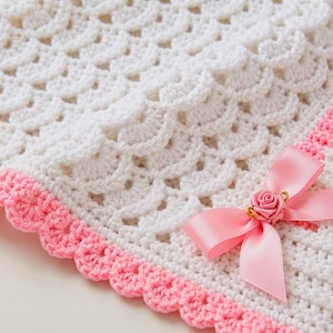 May include: A white crocheted blanket featuring a textured shell stitch pattern throughout. The edges are finished with a scalloped pink crochet border. A pink satin ribbon bow with a small pink fabric rose is attached to the corner.