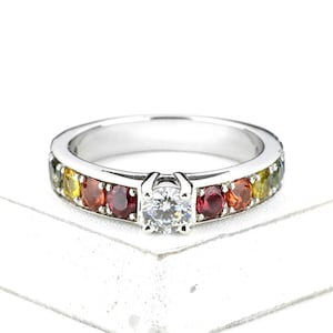 Puede incluir: Un anillo de compromiso de oro blanco con un diamante redondo central y una fila de zafiros arcoíris engastados en la banda.