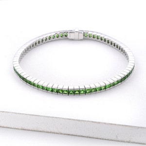 Puede incluir: Una pulsera de plata con una fila de piedras preciosas cuadradas verdes.