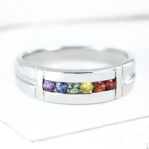 Puede incluir: Un anillo de banda de plata con una fila de piedras preciosas de colores. Las piedras preciosas están engastadas en un engaste de canal y están dispuestas en un patrón de arcoíris. El anillo es de estilo clásico y sería un gran regalo para una ocasión especial.