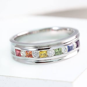 以下が含まれることがあります： 虹色の宝石が並んだシルバーのバンドリング。宝石は正方形にセットされ、虹色のパターンで配置されています。リングは磨かれた仕上げです。