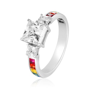 May include: A silver ring with a large square-shaped clear gemstone and two smaller clear gemstones on either side. The band of the ring is decorated with a rainbow of colored gemstones.