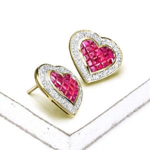 May include: A pair of heart-shaped earrings with a gold setting and a red ruby centre. The earrings are surrounded by a halo of smaller diamonds.