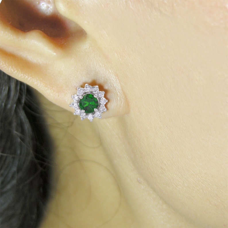 May include: A pair of silver stud earrings with a green gemstone surrounded by a halo of smaller white gemstones.