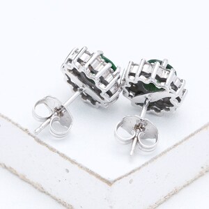 May include: A pair of silver earrings with green gemstones set in a halo of smaller clear stones. The earrings have a post and butterfly back.