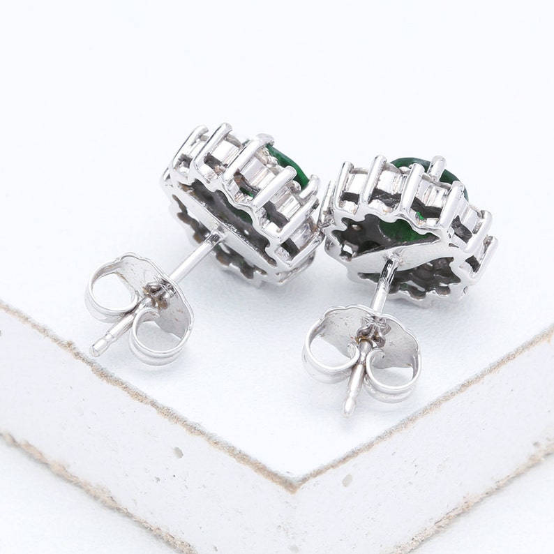 May include: A pair of silver earrings with green gemstones set in a halo of smaller clear stones. The earrings have a post and butterfly back.