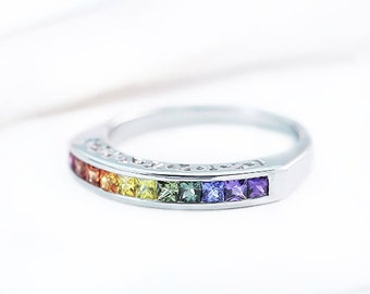 Puede incluir: Un anillo de banda de plata con una fila de piedras preciosas cuadradas de color arcoíris. Las piedras preciosas son rojas, naranjas, amarillas, verdes, azules y moradas.