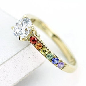 Puede incluir: Un anillo de banda de oro con una piedra central grande, redonda y transparente y un arcoíris de piedras más pequeñas engastadas en la banda.