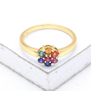 Puede incluir: Un anillo de banda de oro con un racimo de piedras preciosas de colores en el centro. Las piedras preciosas están dispuestas en forma de flor e incluyen rojo, naranja, amarillo, verde, azul y morado.