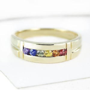 Puede incluir: Un anillo de banda de oro con una fila de piedras preciosas de colores. Las piedras preciosas están engastadas en un engaste de canal y están dispuestas en un patrón de arcoíris. El anillo es de estilo clásico y sería un gran regalo para una ocasión especial.