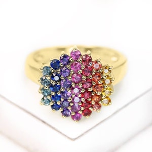 Puede incluir: Un anillo de oro con un gran grupo de piedras preciosas de colores. El grupo de piedras preciosas está dispuesto en forma de arcoíris, con piedras preciosas azules, moradas, rosas, rojas, naranjas y amarillas.