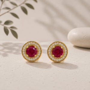 Puede incluir: Un par de pendientes de botón redondos con una piedra preciosa roja circular central rodeada por un halo de pequeñas piedras blancas engastadas en un marco de oro amarillo. Los pendientes presentan un diseño clásico con engaste de garras y borde texturizado.