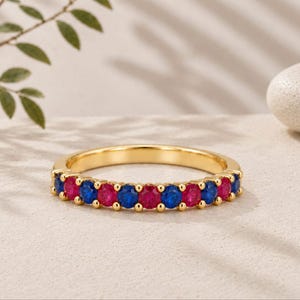 Op de afbeelding: Een gouden ring met een rij afwisselend blauwe en roze ronde edelstenen in een klauwzetting. De ring heeft een gepolijste metalen afwerking en een consistente cirkelvorm, wat het contrast tussen de kleurrijke stenen duidelijk naar voren brengt.