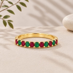 Op de afbeelding: Een gouden ring met een afwisselende rij ronde rode en groene edelstenen in een klauwzetting. De ring toont een doorlopend patroon van levendige karmozijnrode en smaragdgroene stenen langs de voorste helft van de metalen ringband.