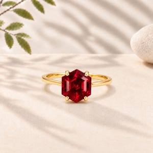 Puede incluir: Un anillo de oro con una prominente piedra preciosa de color rojo intenso tallada en forma hexagonal y montada en seis garras. El aro es liso y pulido, destacando la vibrante piedra carmesí como el punto focal central de esta joya.