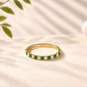 Puede incluir: Un anillo de banda fina de oro con un patrón alterno de piedras redondas transparentes y piedras redondas de color verde vibrante engastadas continuamente alrededor de toda la banda.