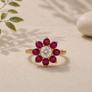 May include: A gold ring featuring a central round white gemstone surrounded by eight oval-cut deep pinkish-red gemstones arranged in a floral pattern. The band is thin and polished gold, creating a bright contrast with the vibrant red and white stones.