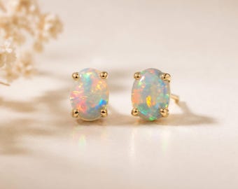 May include: A pair of oval-shaped opal stud earrings set in gold metal prongs. Each opal displays a translucent white base with iridescent flashes of orange, green, and blue light. The earrings feature a classic four-prong setting design.