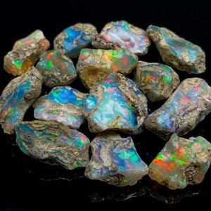 May include: A collection of raw, unpolished opal stones featuring a rough, earthy exterior texture and vibrant, iridescent flashes of blue, green, and red light emanating from within the crystalline structures against a solid black background.