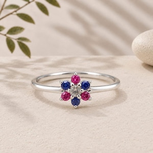 May include: A silver-toned ring featuring a central cluster of six small, round-cut gemstones arranged in a flower-like pattern. The cluster consists of three vibrant pink stones and three deep blue stones surrounding a single clear centre stone.