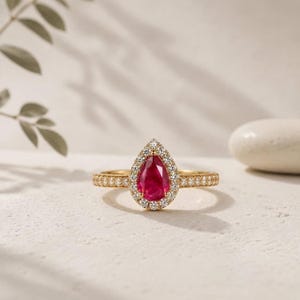 May include: A gold ring featuring a pear-shaped red ruby center stone surrounded by a halo of small white diamonds. The band is also set with a row of small white diamonds extending along the sides, creating a bright and elegant appearance.