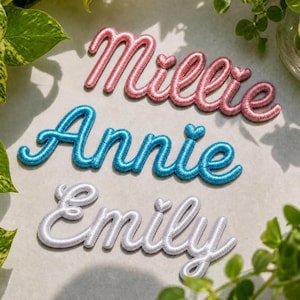 May include: Three embroidered name patches in cursive script. The top patch is pink and reads Millie, the middle patch is blue and reads Annie, and the bottom patch is white and reads Emily. Each name features a small heart above the letter i.