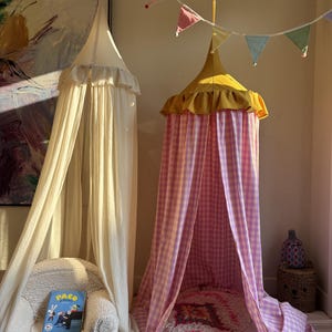 May include: Two fabric canopies hanging from the ceiling. One is solid cream with a ruffled top, positioned over a white textured armchair with a book titled Paco. The other is a yellow and pink gingham canopy with a yellow ruffled top.