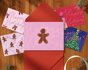May include: A set of five rectangular greeting cards featuring holiday themes, including a gingerbread man, a Christmas tree, and bows, with the text Happy Holidays written in cursive script, displayed with a red envelope on a wooden surface.