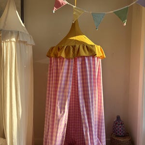 May include: A hanging canopy featuring a mustard yellow conical top with a ruffled edge and long, flowing panels of pink and white gingham fabric. A string of pastel-colored triangular bunting hangs above the canopy, adding a decorative touch.