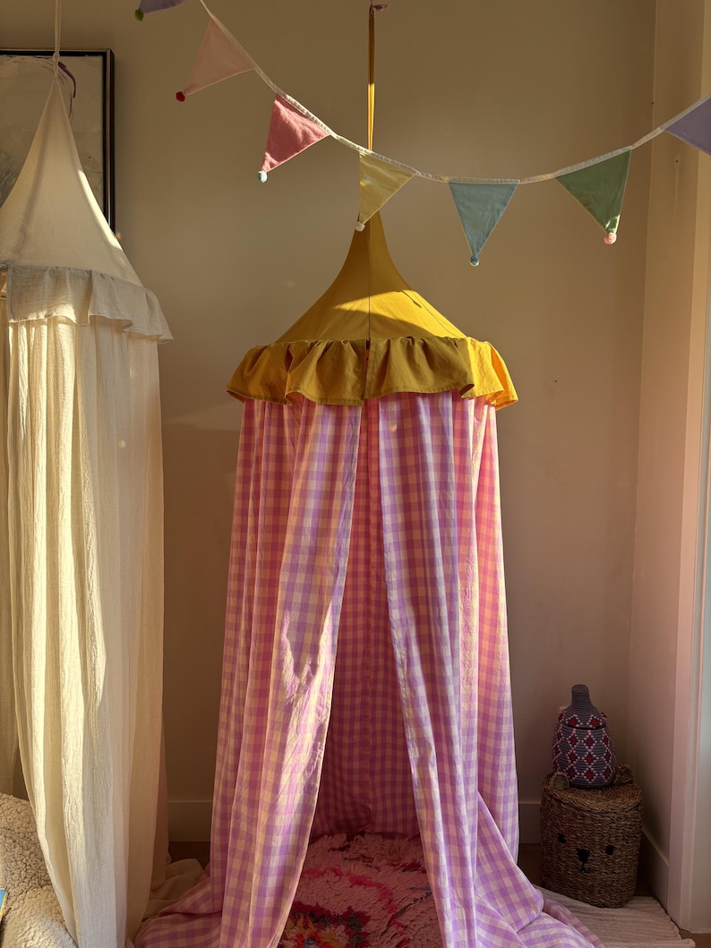 May include: A hanging canopy featuring a mustard yellow conical top with a ruffled edge and long, flowing panels of pink and white gingham fabric. A string of pastel-colored triangular bunting hangs above the canopy, adding a decorative touch.