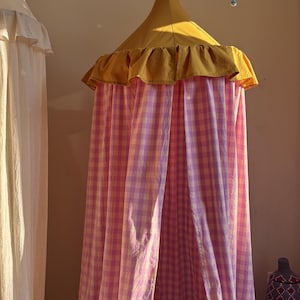 May include: A hanging canopy featuring a mustard yellow conical top with a ruffled edge and long, flowing curtains in a pink and white gingham check pattern. A string of pastel-colored triangular fabric bunting hangs above the canopy structure.
