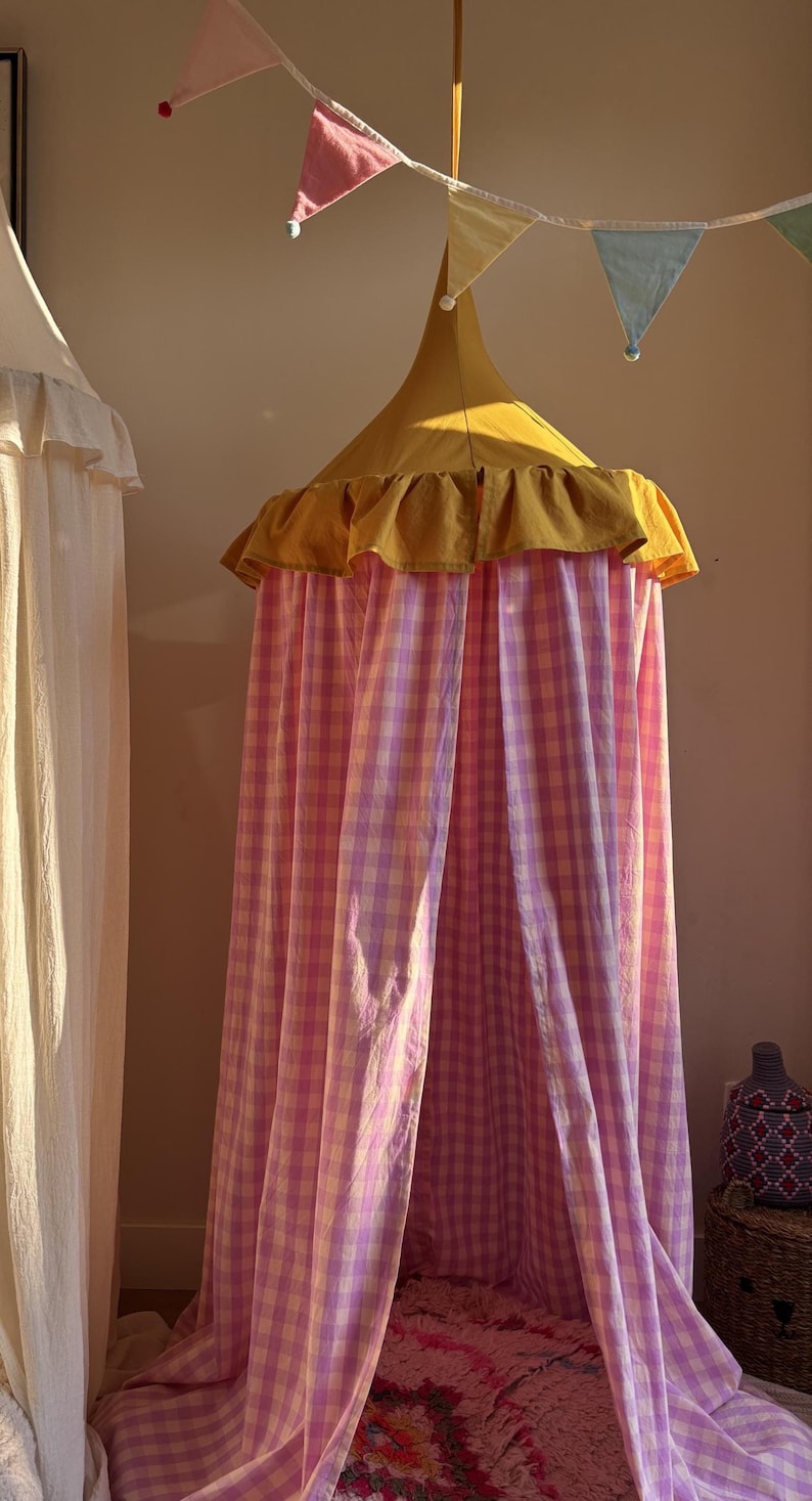 May include: A hanging canopy featuring a mustard yellow conical top with a ruffled edge and long, flowing curtains in a pink and white gingham check pattern. A string of pastel-colored triangular fabric bunting hangs above the canopy structure.