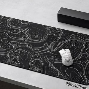 May include: A rectangular black desk mat featuring a white topographic map line pattern. The mat measures 900x400mm and is accompanied by a white computer mouse with a honeycomb shell design, a black rectangular storage box, and a rolled-up mat.