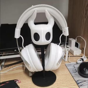 Peut inclure: Un support de casque blanc représentant une tête de personnage stylisée avec deux cornes incurvées vers le haut et deux grands yeux ovales noirs. Le support possède un mât vertical noir et une base circulaire noire pour un casque audio.
