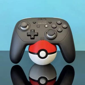 Puede incluir: Un mando Nintendo Switch Pro negro apoyado sobre un soporte esférico rojo y blanco con forma de Poké Ball. El mando cuenta con una cruceta, dos joysticks analógicos y botones etiquetados como A, B, X e Y. El logotipo de Nintendo Switch es blanco.