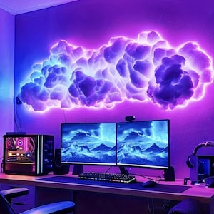 May include: A large, wall-mounted cloud light fixture emitting a vibrant purple and blue glow. The cloud-shaped structure features textured, fluffy layers illuminated from within, positioned above a dual-monitor computer setup on a desk.