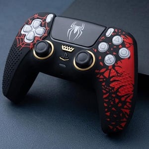 Peut inclure: Une manette de jeu vidéo noire avec un motif de toile d'araignée rouge sur les poignées, un emblème d'araignée blanc sur le pavé tactile et une icône de couronne dorée au-dessus du logo PlayStation central, avec des boutons argentés.