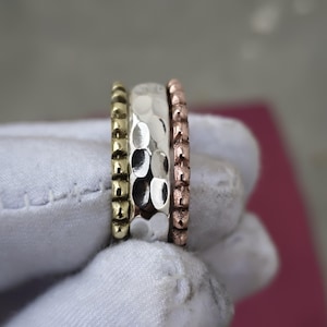 May include: A set of three stacked rings featuring a gold-toned band with a beaded texture, a central silver-toned band with a hammered finish, and a copper-toned band with a textured surface, all displayed together in a vertical arrangement.