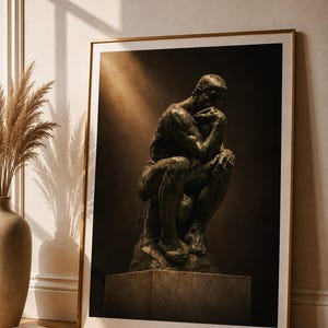 Puede incluir: Una impresión de arte mural enmarcada que muestra una escultura de tono bronce oscuro de una figura sentada en pose contemplativa, con la barbilla apoyada en una mano. La obra está en un marco fino de color dorado, con un juego de luces y sombras.